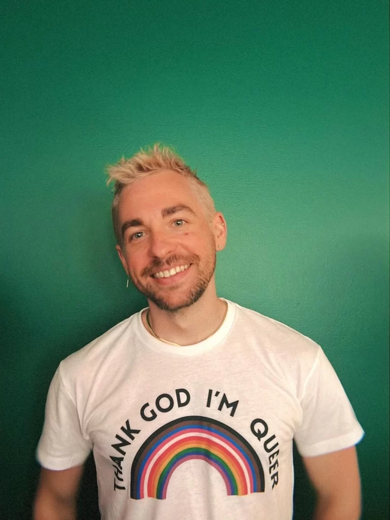 Ian Helms stands in front of a green wall, smiling with bleached blonde hair and wearing a white "thank god i'm queer" shirt with an LGBTQ+ rainbow beneath the text
